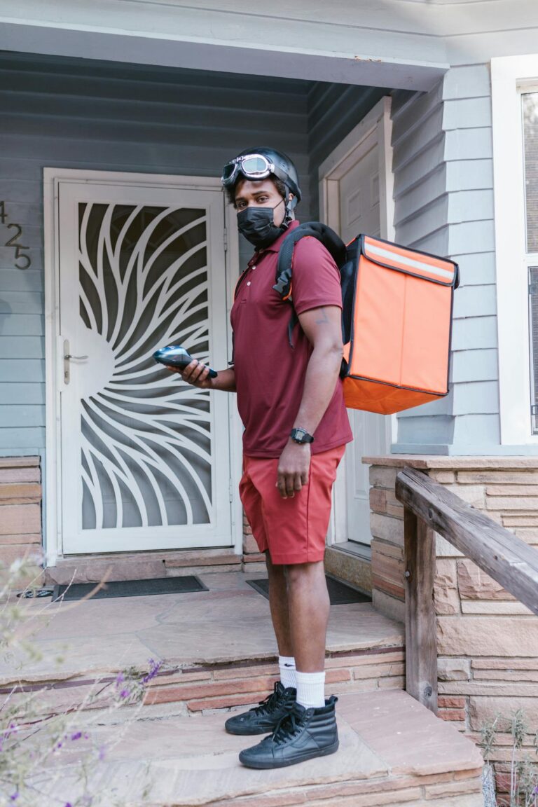 Courier with delivery bag and helmet waits at a porch, ready for shipment.